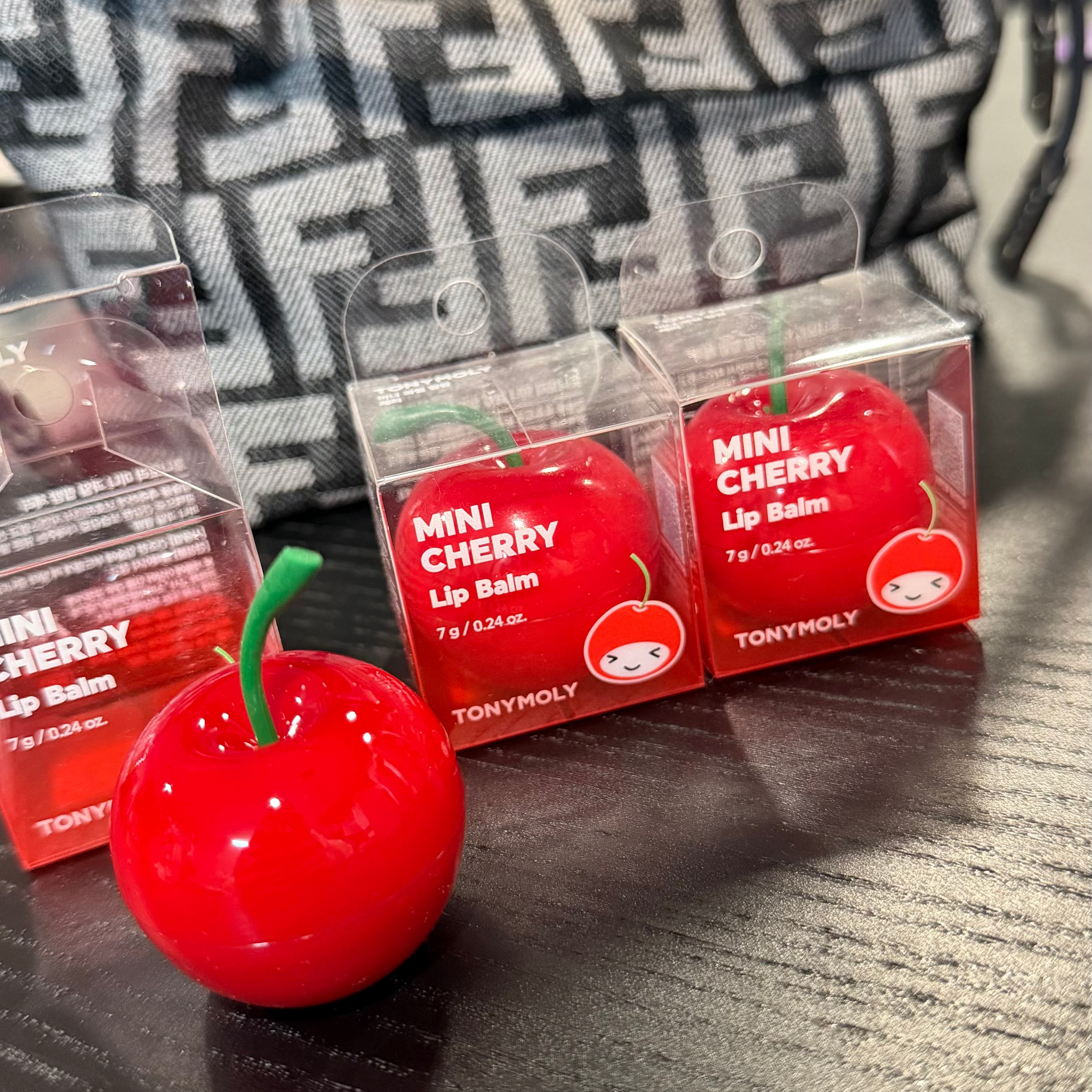 A glossy red cherry-shaped lip balm with a green stem sits on a dark wooden table in front of its clear packaging boxes labeled 'MINI CHERRY Lip Balm' by TONYHOLY. A black and white patterned bag is blurred in the background.